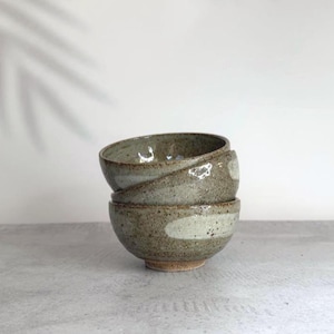 May include: A stack of two ceramic bowls with a speckled, earthy brown and grey glaze. The bowls feature a smooth, lighter-coloured horizontal band across the exterior surface, showcasing a rounded shape and a visible footed base at the bottom.