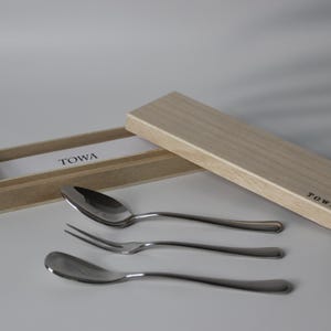 Puede incluir: Un juego de cubiertos de acero inoxidable con una cuchara, un tenedor y dos cucharas, junto con una caja de madera. La caja está abierta, revelando una etiqueta blanca con la palabra "TOWA". La tapa de la caja también es visible, con la palabra "TOWA" impresa.