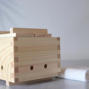 May include: A rectangular light-coloured wooden tofu press featuring visible wood grain, finger-jointed corners, and four circular drainage holes along the side. A flat wooden lid rests on top, and a folded white cloth is placed beside the press.