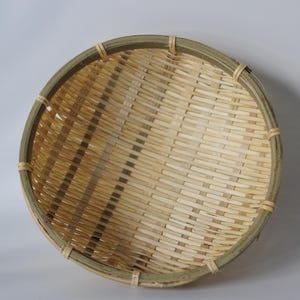 May include: A round, shallow basket constructed from woven light-colored bamboo strips. The basket features a reinforced, darker green-toned bamboo rim secured with thin, light-colored bamboo bindings at regular intervals around the circular edge.
