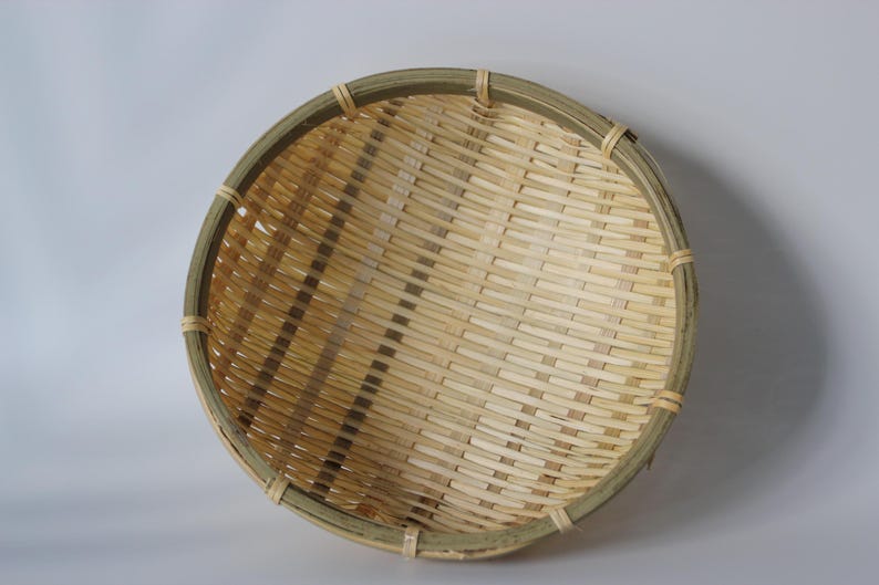 May include: A round, shallow basket constructed from woven light-colored bamboo strips. The basket features a reinforced, darker green-toned bamboo rim secured with thin, light-colored bamboo bindings at regular intervals around the circular edge.