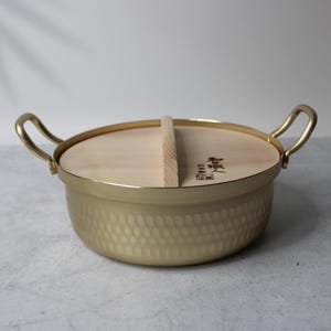 May include: A gold-coloured, hammered metal cooking pot with a wooden lid. The pot has two curved handles and a wooden lid with a vertical handle. The lid has Japanese text. The pot is designed for cooking and serving.