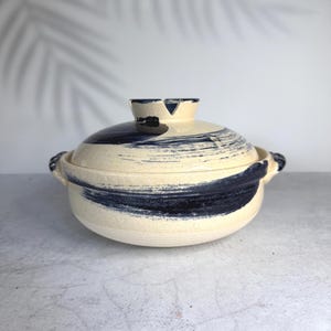 Puede incluir: Una cazuela de cerámica redonda con tapa a juego, que presenta una base de color crema con patrones de pinceladas azul oscuro. La olla incluye dos pequeñas asas laterales y un pomo central en la tapa con un diseño de muesca.