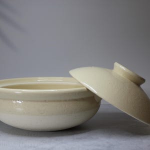 Peut inclure: Pot de cuisson en c&eacute;ramique cr&egrave;me avec un couvercle assorti. Le pot a une forme arrondie avec deux petites poign&eacute;es et une large ouverture. Le couvercle est l&eacute;g&egrave;rement entrouvert.