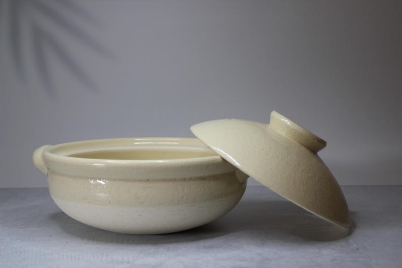 Peut inclure: Pot de cuisson en c&eacute;ramique cr&egrave;me avec un couvercle assorti. Le pot a une forme arrondie avec deux petites poign&eacute;es et une large ouverture. Le couvercle est l&eacute;g&egrave;rement entrouvert.