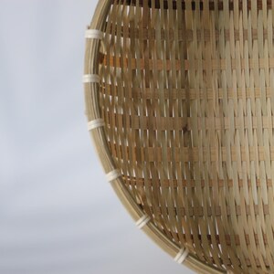 May include: A round, shallow basket constructed from light tan woven bamboo strips. The rim is reinforced with a thicker bamboo band secured by evenly spaced white cord bindings, creating a structured, open-weave pattern throughout the body.