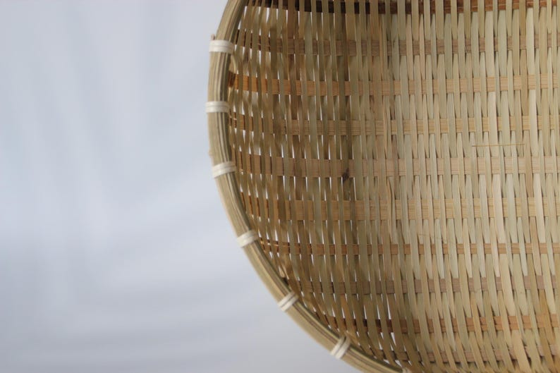 May include: A round, shallow basket constructed from light tan woven bamboo strips. The rim is reinforced with a thicker bamboo band secured by evenly spaced white cord bindings, creating a structured, open-weave pattern throughout the body.