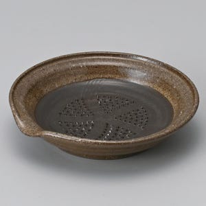 May include: A brown ceramic grater with a dark interior. The grater has a circular shape with a slightly raised rim and a small spout. The interior features a pattern of small, triangular grating surfaces.
