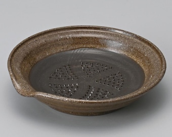 May include: A brown ceramic grater with a dark interior. The grater has a circular shape with a slightly raised rim and a small spout. The interior features a pattern of small, triangular grating surfaces.