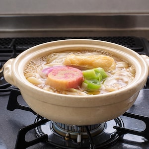 Peut inclure: Un pot en c&eacute;ramique beige rempli de nouilles udon japonaises, de bouillon et de garnitures vari&eacute;es. Les garnitures comprennent un g&acirc;teau de poisson rose et blanc, des oignons verts et du tofu frit. Le pot est sur une cuisini&egrave;re &agrave; gaz.