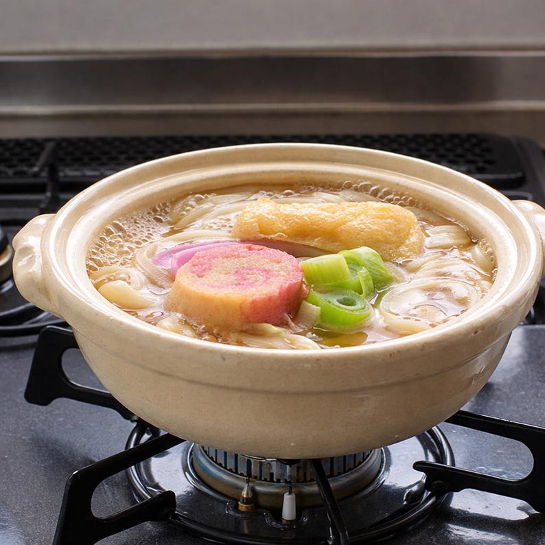 Peut inclure: Un pot en c&eacute;ramique beige rempli de nouilles udon japonaises, de bouillon et de garnitures vari&eacute;es. Les garnitures comprennent un g&acirc;teau de poisson rose et blanc, des oignons verts et du tofu frit. Le pot est sur une cuisini&egrave;re &agrave; gaz.