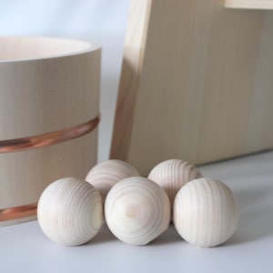 Op de afbeelding: Een lichtgekleurde houten emmer met twee horizontale koperen banden rond de buitenkant, vergezeld van vijf gladde, bolvormige houten ballen gerangschikt in een groep voor de emmer en een plat houten bord erachter geplaatst.