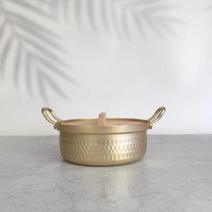 Puede incluir: Una olla redonda de metal dorado con una textura martillada en el exterior. Cuenta con dos asas curvas en los lados opuestos y una tapa circular plana con un pequeño mango de madera de color claro centrado en la parte superior.