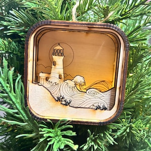 May include: A wooden ornament featuring a lighthouse with waves crashing against the shore. The ornament is a light brown color and has a dark brown border.