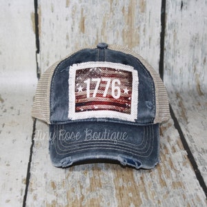 May include: A blue and beige trucker hat with a distressed look. The hat has a white patch with a red, white, and blue American flag design and the number "1776" in white.