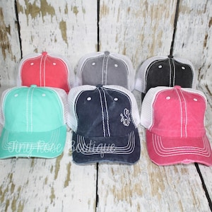 May include: Six different colored trucker hats with white mesh backs. The hats are made of a distressed cotton fabric and have a white stitched design. The hats are arranged in a row on a white wooden surface.