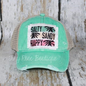 May include: A turquoise and beige distressed trucker hat with a patch that reads "Salty Sandy Happy" with palm trees.