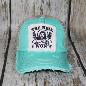 May include: A turquoise baseball cap with a distressed brim and a mesh back. A white rectangular patch is centered on the front, featuring a black graphic illustration of a person and the text THE HELL I WON'T in bold black capital letters.
