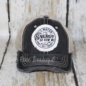May include: A black and beige mesh trucker hat with a distressed look. The hat has a white circle patch with the text "I Match Energy So How We Don't Act Today?" in black.