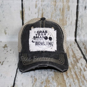 May include: Black and tan distressed trucker hat with a white patch that says "Grab your balls we're going bowling" with a bowling pin and bowling ball graphic.