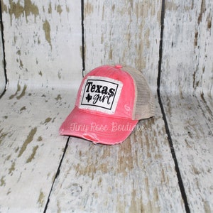 Pu&ograve; includere: Un cappello da camionista rosa e beige rovinato con una toppa bianca che dice "Texas girl" e un contorno nero dello stato del Texas.
