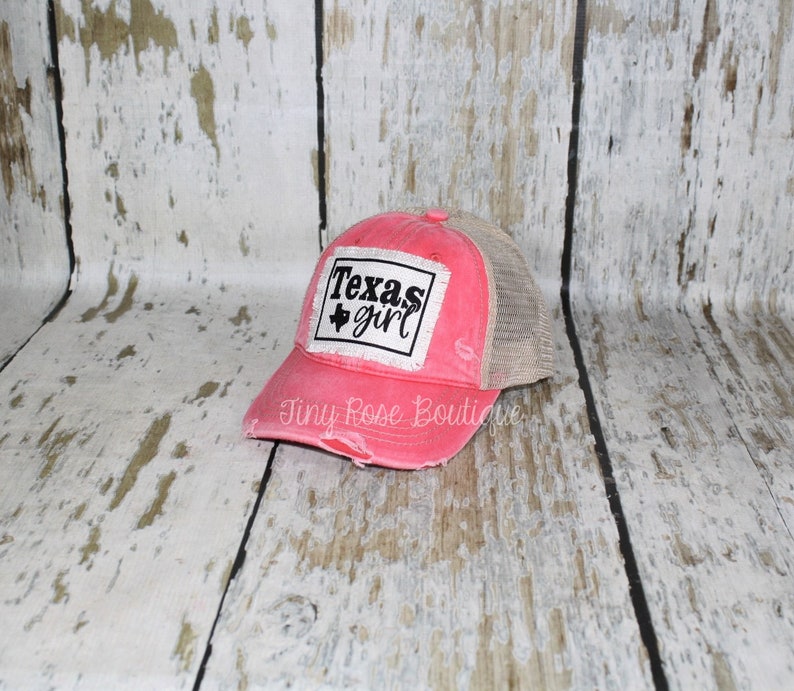 Pu&ograve; includere: Un cappello da camionista rosa e beige rovinato con una toppa bianca che dice "Texas girl" e un contorno nero dello stato del Texas.