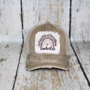 May include: A brown and beige distressed trucker hat with a rainbow patch that says "teacherlife" in black text. The rainbow is made of leopard print and has a pink heart in the center.