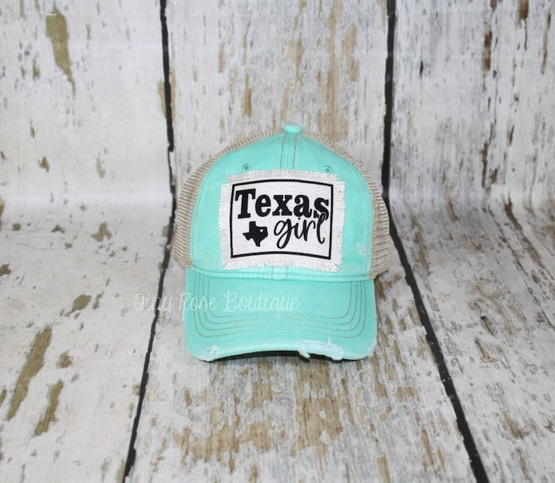 Pu&ograve; includere: Un cappello da camionista turchese e beige dall'aspetto usurato. Il cappello ha una toppa bianca con testo nero che recita "Texas girl" e un piccolo contorno nero dello stato del Texas.