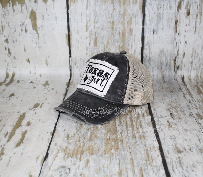 Pu&ograve; includere: Cappellino da camionista nero e beige usurato con una toppa bianca che dice "Texas girl" in lettere nere.