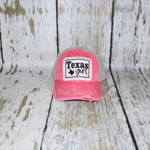 Pu&ograve; includere: Un cappello da camionista rosa e beige consumato con una toppa nera che dice "Texas girl" e un contorno nero dello stato del Texas.
