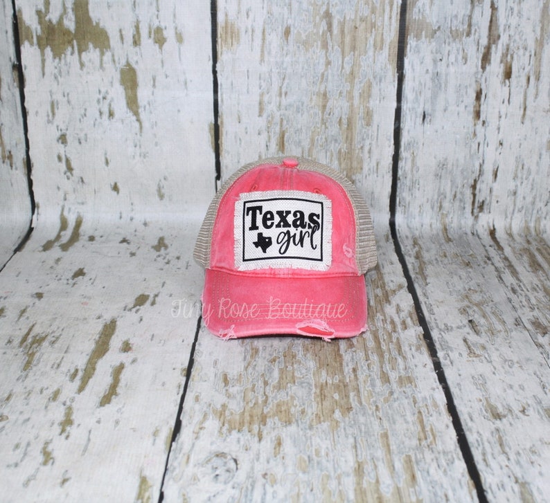 Pu&ograve; includere: Un cappello da camionista rosa e beige consumato con una toppa nera che dice "Texas girl" e un contorno nero dello stato del Texas.