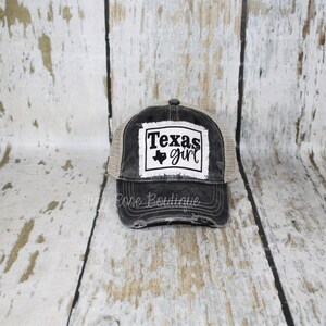 Pu&ograve; includere: Cappellino da camionista nero e marrone rovinato con una toppa bianca che dice "Texas girl" e un contorno nero dello stato del Texas con un cuore al centro.