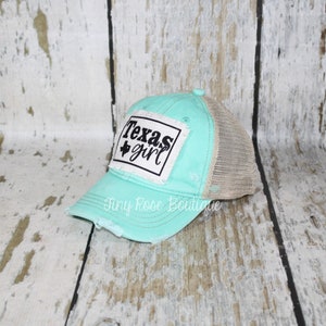 Pu&ograve; includere: Un cappello da camionista in rete turchese e beige con un aspetto usurato. Il cappello ha una toppa bianca con testo nero che recita "Texas girl" e una piccola stella nera.