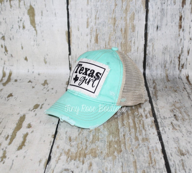 Pu&ograve; includere: Un cappello da camionista in rete turchese e beige con un aspetto usurato. Il cappello ha una toppa bianca con testo nero che recita "Texas girl" e una piccola stella nera.