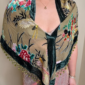 Op de afbeelding: Een olijfgroene sjaal met een bloemenpatroon in rood, roze en blauw. De sjaal heeft een donkergroene fluwelen rand en is versierd met groene kralenfranjes. Een gouden ketting is zichtbaar.