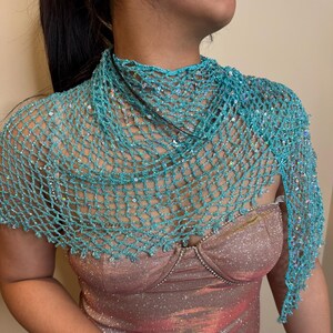 May include: A turquoise crocheted shawl with iridescent sequins. The open-weave design drapes over the shoulders. The shawl is a delicate accessory, suitable for special occasions or adding a touch of elegance to an outfit.
