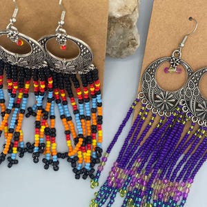 May include: Two pairs of beaded earrings. One pair features silver-toned oval pendants with black, red, orange, yellow, and blue beads. The other pair has silver-toned pendants with purple, green, and yellow beads. Both pairs have decorative metalwork.