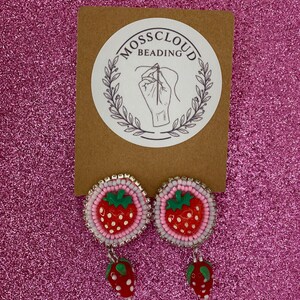 May include: A pair of beaded strawberry earrings. Each earring features a red strawberry design with green leaves and white dots. The earrings are attached to a pink and white beaded circle, with a silver-colored border. The earrings are displayed on a brown card with the text "Mosscloud Beading".