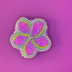 May include: A pink, green, and yellow beaded flower with a clear rhinestone center. The flower is on a purple background.