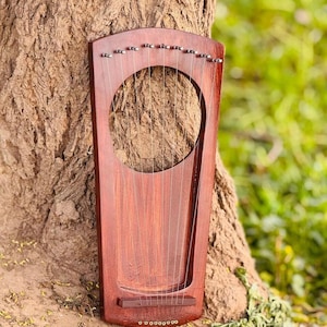 May include: A dark reddish-brown wooden lyre featuring a large circular sound hole in the upper section, ten thin metal strings stretched vertically across the body, and ten small silver tuning pegs positioned along the top edge of the instrument.