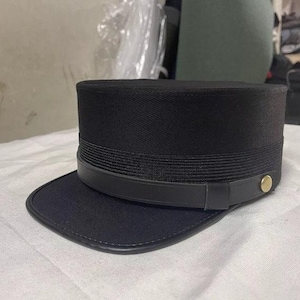 May include: A black, flat-topped hat with a black leather band and a gold button. The hat has a wide brim.