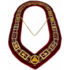 Puede incluir: Un collar de terciopelo rojo con eslabones rectangulares dorados y emblemas decorativos. Una cadena dorada conecta los dos extremos. El collar está diseñado con varias formas y símbolos geométricos, probablemente para fines ceremoniales.