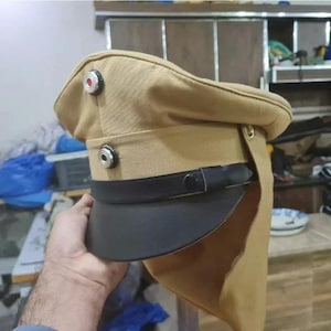 Pode incluir: Chapéu de estilo militar bege com viseira preta e correia de couro preta. O chapéu tem dois botões prateados na frente e um botão vermelho e prateado na parte superior. O chapéu é feito de tecido bege.