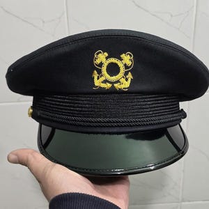 Könnte beinhalten: Schwarze Mütze im Militärstil mit glänzendem, dunkelgrünem Visier. Der Hut hat ein goldenes Emblem und Zierleisten, mit einem schwarzen Band um die Krone. Auf der Seite ist ein goldener Knopf zu sehen. Der Hut wird von einer Hand gehalten.