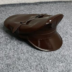 以下が含まれることがあります： 光沢のあるパテントレザーのような仕上げのダークブラウンのキャップ。この帽子は、構造化された丸みを帯びたクラウン、硬いカーブしたバイザー、そしてバイザーの上の前面を水平に包む装飾的なストラップのディテールが特徴です。