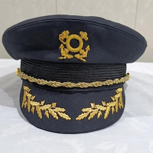 May include: Navy blue hat with gold embroidered details. The hat features a gold emblem on the front and gold leaf designs on the brim. A gold rope trim encircles the hat. The hat is likely a uniform hat.