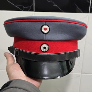 Puede incluir: Un sombrero de estilo militar gris y rojo con visera negra. El sombrero presenta una banda y ribete rojos, con dos botones circulares en la parte delantera. El sombrero es sostenido por una mano.