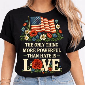 Può includere: T-shirt nero con un design della bandiera americana e fiori rossi, bianchi e blu. Il testo recita: "THE ONLY THING MORE POWERFUL THAN HATE IS LOVE."