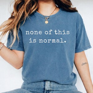 May include: A blue t-shirt with the white text "none of this is normal". The shirt has a crew neck and short sleeves. The model is wearing blue jeans and a gold necklace.
