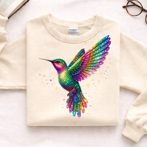 Op de afbeelding: Crèmekleurige sweatshirt met een kleurrijk kolibrie-ontwerp. De vogel heeft iriserende vleugels in groen, roze, paars en geel. Het ontwerp bevat kleine stervormige accenten.